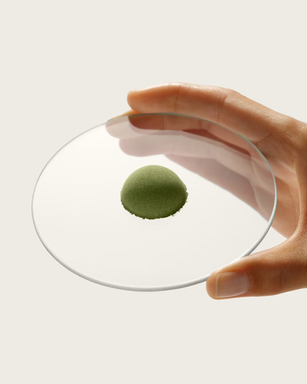 A hand holding a clear watch-glass with a small mound of green Alutic powder, in soft directional studio light against a warm neutral background.