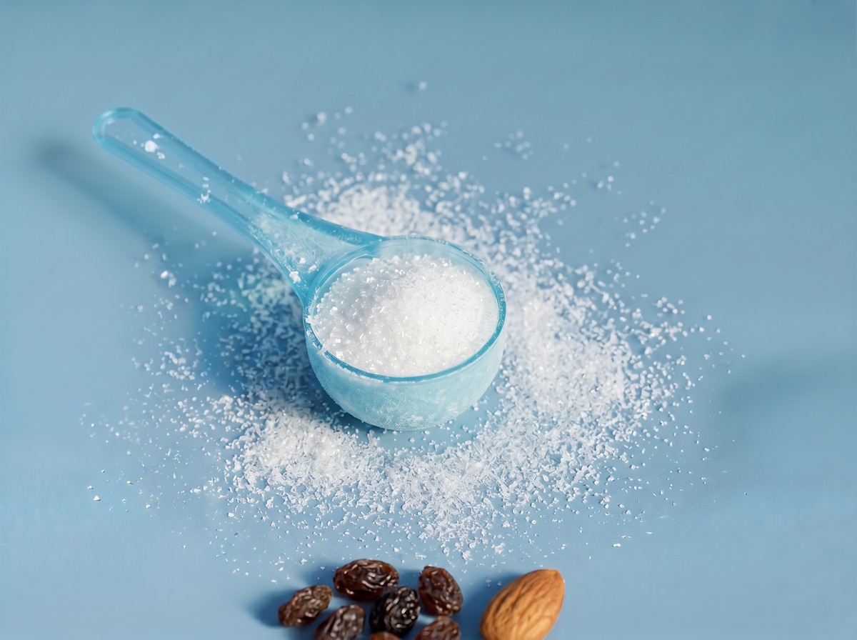 A translucent blue scoop of crystalline white boron powder with raisins and an almond at the bottom edge on a muted blue studio background.