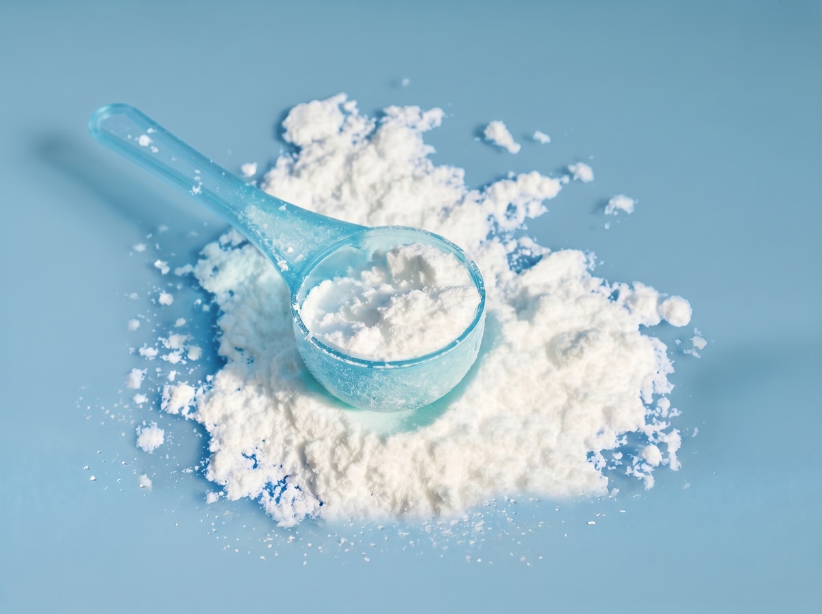 A translucent blue scoop overflowing with fine white creatine monohydrate powder on a muted blue studio background.