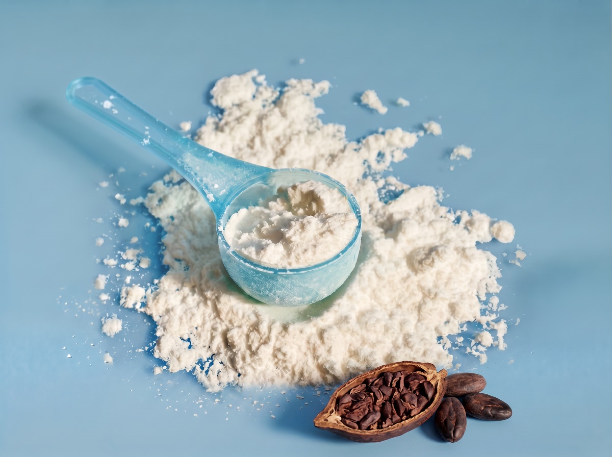 A translucent blue scoop of off-white epicatechin flavanol extract powder with a halved cacao bean and whole beans at the bottom edge on a muted blue studio background.