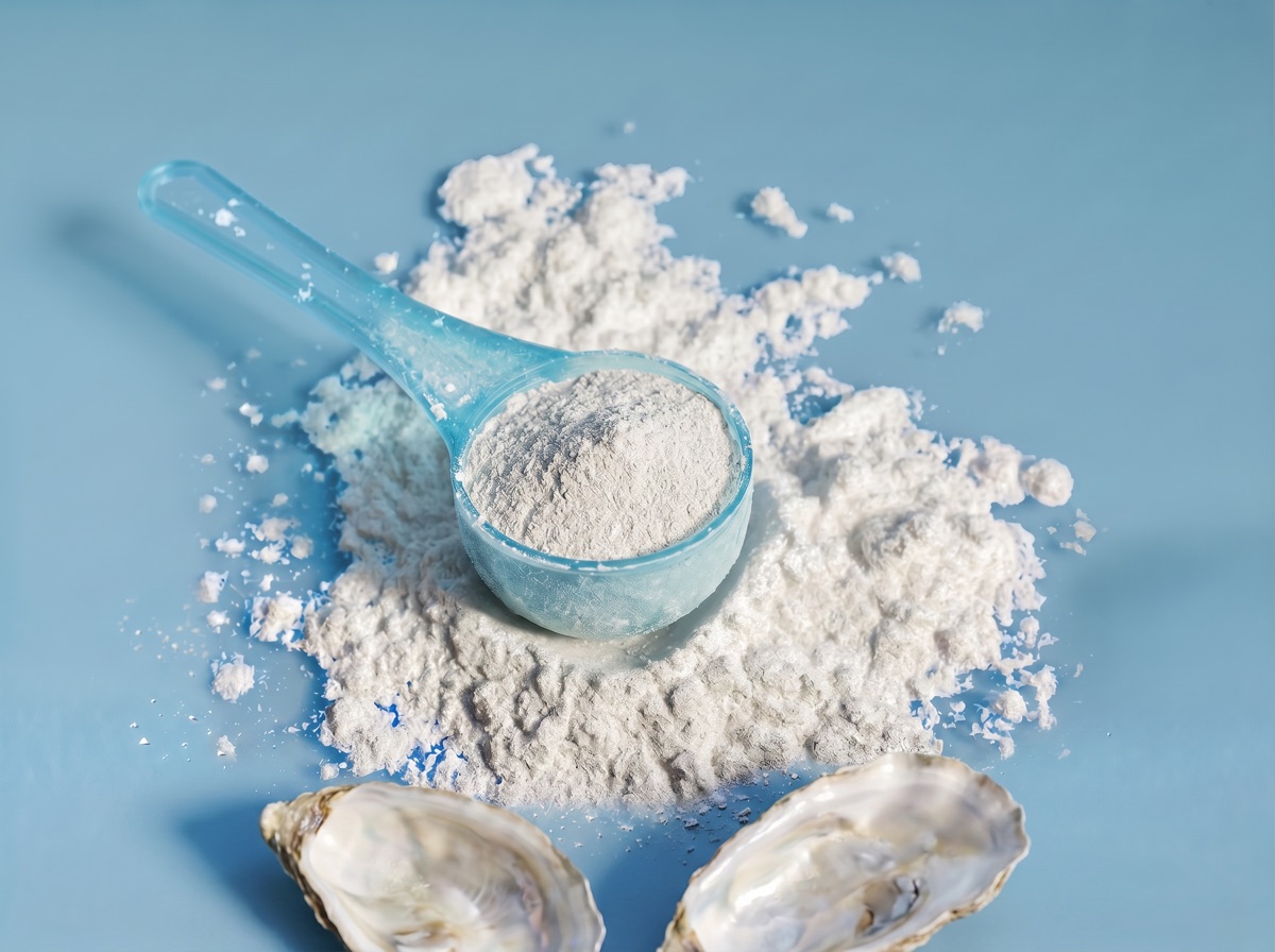 A translucent blue scoop of pale grey-white zinc powder with two opened oyster shells at the bottom edge on a muted blue studio background.