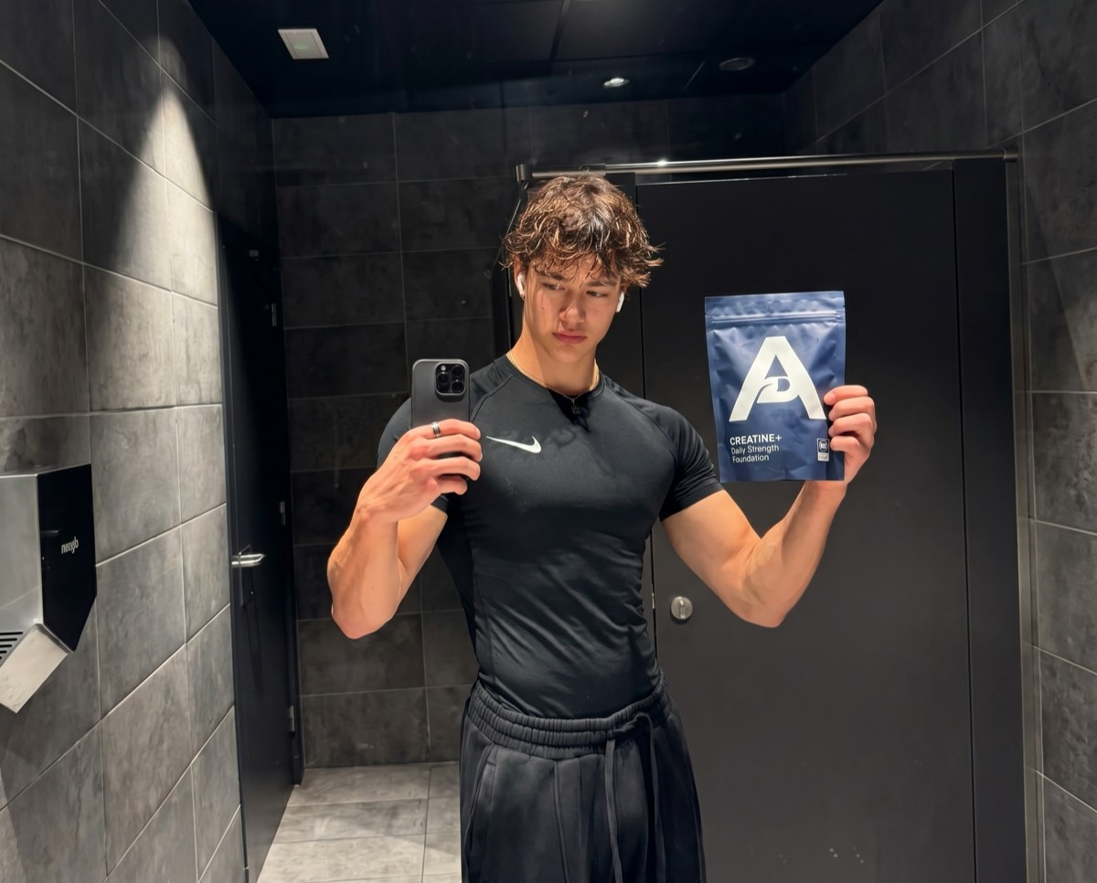 A customer in a gym taking a mirror selfie holding the Alutic Creatine+ pouch after a training session.