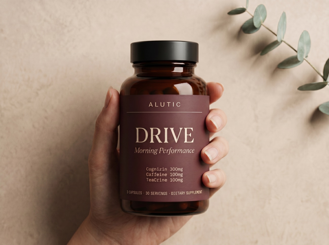 A hand holding the Alutic Drive bottle against a warm neutral wall with a eucalyptus sprig.